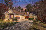 A cozy, single-story house with stone and wood siding, gabled roof, and warm exterior lights sits at 114 Blue Heron Drive in Youngsville, beside a wooded area at sunset. A concrete driveway leads to the garage and front entrance.