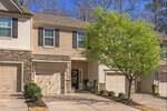 A two-story townhouse at 1434 Chatuga Way, Wake Forest, with tan siding, stone accents, black shutters, and a garage. A leafy green tree stands in front, casting shade on the bright and well-maintained driveway and entrance.