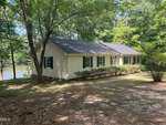 Nestled at 113 Country Brook Lane in Youngsville, this single-story house with light siding and green shutters sits beside a tranquil lake, surrounded by trees and bushes with sunlight filtering through the leaves.