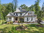 A large, modern two-story white house with dark roof shingles at 3511 Bruce Garner Road in Wake Forest features multiple gables, a spacious porch, a green lawn, landscaped garden with a circular stone fountain, and is surrounded by tall trees.