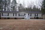 A single-story, gray manufactured home with black shutters and white railings sits on a large, brown lawn at 70 Cole Circle in Franklinton, surrounded by leafless trees and pine trees under a cloudy sky.