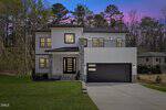 Modern two-story house at 102 Horseman Drive, Louisburg, featuring white vertical siding, large black-trimmed windows, a double garage, stone accents, and a balcony. Trees and a purple sunset sky create a picturesque backdrop.
