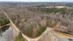 Aerial view of 5590 Shelton Creek Road, Oxford, with intersecting dirt roads, leafless trees, a small pond to the left, scattered houses, and forested land stretching into the distance beneath a cloudy sky.