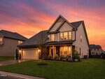 A modern two-story suburban house at 405 Hickory Run Lane, Youngsville, with a well-kept lawn and illuminated windows at dusk. A colorful sunset sky rises behind shrubs and flowers that decorate the front yard.