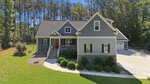 Nestled in Louisburg, 142 Mohawk Drive is a gray single-family house with white trim, a front porch, and red door. Set before a wooded area, it features neat landscaping, shrubs along the walkway, and a driveway to a detached garage.