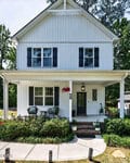 A two-story white house with black shutters and a covered front porch at 5192 NC 56 Hwy, Franklinton, featuring outdoor seating, potted plants, and a "HOME" sign above the door. Green shrubs and a curved walkway complete the inviting look.