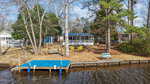 A small lakeside cabin at 1607 Sagamore Drive, Louisburg, with a screened porch, sits among tall trees. In front are yellow Adirondack chairs, a wooden swing, and a blue bench. A dock with a blue cover stretches into the water.