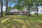 A brick house with a white porch sits behind large trees on a grassy lawn at 3916 US 401 Highway N, Louisburg, with shrubs along the front and a partly cloudy blue sky in the background.