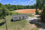 A small white manufactured home sits on a grassy lot at 4004 Old NC 75 in Stem, with a gravel driveway, surrounded by trees and next to freshly plowed farmland under a blue sky with scattered clouds.