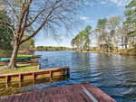 A wooden dock extends into a peaceful lake surrounded by trees and houses under a partly cloudy blue sky at 959 Sagamore Drive, Louisburg. Canoes are visible on the grassy bank to the left.