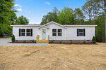 Charming single-story white house with black shutters at 105 Buck Ridge Road, Louisburg. Features a small front porch with steps, gravel pathway, and is surrounded by trees and straw-covered ground.