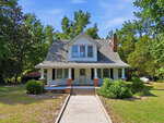 A white two-story house at 439 Lake View Road, Louisburg, features a large front porch with brick steps and columns, surrounded by trees and greenery under a clear blue sky. A concrete path leads up to the entrance.