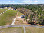 Aerial view of a single-story house at 1027 Johnson Town Road, Zebulon, with a long driveway, surrounded by grass and trees in a spacious rural neighborhood near a main road with several other distant homes.