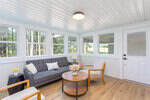 A bright sunroom with large windows, a gray sofa, two wooden armchairs, a round coffee table, and a white door. The room has white walls and ceiling, wood flooring, and simple modern decor.