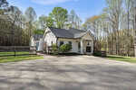 Nestled next to a wooded area in Youngsville, 2566 Mays Crossroad Road features a white single-story house with a dark roof, surrounded by trees and shrubs; a paved driveway leads to the entrance, and a wooden fence lines the yard.