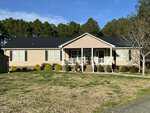 Nestled at 1195 US 158 in Oxford, this single-story beige house features a black roof, white trim, and a covered front porch with steps. The yard boasts patchy grass and small shrubs, bordered by tall pines under a blue sky.