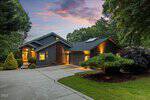A modern house at 605 Sagamore Drive, Louisburg, features a dark exterior and warm lighting, nestled among trees at sunset. A curved concrete driveway leads to a two-car garage, with manicured landscaping enhancing the serene atmosphere.