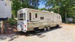A white Outback travel trailer with two axles is parked on a gravel lot at 219 Eagle Drive in Louisburg, next to a white house and surrounded by trees and greenery on a sunny day.