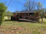 A single-story red brick house with a black roof, front porch, and ramp at 214 E Persimmon Street, Youngsville, surrounded by a grassy yard, trees, and bushes under a clear blue sky.