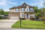 Charming two-story beige house at 25 Cranbrooke Drive, Franklinton, features stone accents, a blue front door, double garage, front porch, manicured lawn, mailbox at the curb, driveway, and trees and fence under a blue sky.