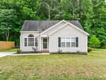 A small, single-story gray house with white trim, black shutters, and a front porch sits at 65 Meadowfield Lane in Franklinton. An American flag hangs by the door, surrounded by grass, trees, and a wooden fence on the left.