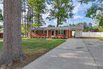 Charming single-story brick house at 114 Edgewood Drive, Louisburg, with white trim, a covered carport, and spacious driveway. Enjoy a lush yard with green grass, trees, and a white privacy fence under a blue sky with scattered clouds.