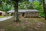 A single-story brick house at 479 Ridgecrest Trail, Henderson, features large front windows, a covered carport on the left, and a spacious yard shaded by tall trees. The mostly bare lawn has patches of grass and the home is surrounded by lush greenery.