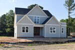 Newly constructed two-story house at 215 Broadleaf Lane in Youngsville, featuring gray vertical siding, white trim, multiple gables, and a covered front porch. The yard is unfinished with exposed soil and construction materials; trees line the background.