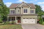 This two-story suburban house at 104 Bonterra Drive, Youngsville, features tan siding, white trim, a double garage, small front porch, manicured shrubs, and a concrete driveway on a green lawn with trees in the background.