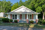 A small, single-story white house with a covered front porch at 50 Robert Sledge Road in Spring Hope. Red shutters, brick columns, and lush greenery surround it on a sunny day, with a detached shed visible in the background.