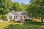 Charming single-story house at 131 N Cobble Creek Drive in Henderson with light-colored siding, a front porch, green shutters, and an American flag. The home is surrounded by a well-kept lawn, small shrubs, and large trees under a clear sky.
