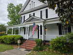 A white, two-story house with black shutters and a covered front porch sits at 401 N Main Street in Louisburg. An American flag hangs near the steps, bordered by green bushes. The yard features trees and a sidewalk.