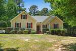 A beige, single-story house at 195 Lumberjack Lane in Louisburg with red shutters and a central front door, surrounded by neatly trimmed bushes and trees. The home sits on a grassy lawn with a wooded backdrop under a blue sky.