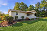 A small, single-story beige house at 1795 Hicks Road, Youngsville, features a front porch with white railing, neatly trimmed shrubs, a well-maintained lawn, and is surrounded by trees under a blue sky.