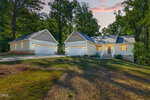 Single-story house at 111 Morning Sun Drive, Louisburg, with two large detached garages, surrounded by trees. Cream siding and gray roofs complement the dusk setting, as warm lights glow from the windows.