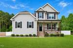 A modern two-story house at 810 Mulberry Road in Spring Hope features gray siding, black shutters, and a covered front porch. The well-manicured lawn with small shrubs is set against a backdrop of trees and a bright blue sky.