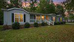 A single-story blue house with white trim and black shutters on 1635 US 15 in Creedmoor, featuring a front porch with white railings, surrounded by green shrubbery and trees, photographed at dusk with warm lights glowing from the windows.