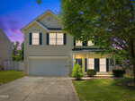 A two-story beige house with dark shutters at 841 Stackhurst Way, Wake Forest, featuring a double garage and covered front porch. Lit from inside, it sits on a green yard with a tree in front, photographed at dusk beneath a purple-blue sky.