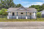 A small white house with blue trim and a gray roof sits behind a black chain-link fence on quiet 1311 S William Street in Henderson. A covered front porch welcomes you, with trees and greenery visible in the background.