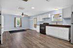 Open-concept kitchen and living area with white cabinets, dark countertops, wood flooring, a ceiling fan, and multiple windows letting in natural light. A black stove and fridge are visible, and the walls are painted light blue.