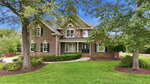 A two-story brick house at 1257 Pine Ridge Road in Zebulon, with black shutters and a black front door, sits among green grass, shrubs, and tall trees beneath a blue sky. A curved walkway leads to the inviting entrance.