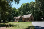 A single-story brick house with white trim and a front porch sits on a large, grassy yard at 946 Cooke Road, Louisburg. Tall trees surround the home, a paved driveway leads up to it, and a basketball hoop is visible on the right.