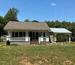 A small white house with a covered front porch and an attached carport sits on a grassy lot at 8549 Grassy Creek Road, Oxford, surrounded by trees and under a clear blue sky.