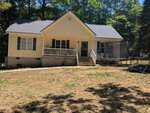 A small yellow house at 172 Nashua Drive, Louisburg, features a covered front porch, green shutters, and a gray roof. The patchy yard hosts a trampoline and outdoor items to the right, with trees surrounding the property.