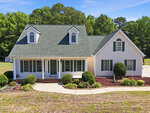 A white, two-story house at 120 Williamston Ridge Drive in Youngsville features green roof shingles, dormer windows, and a front porch. The yard has neatly trimmed bushes, a curved walkway, and is surrounded by trees under a clear blue sky.