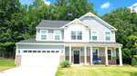 Located at 395 Oak Park Boulevard in Youngsville, this two-story suburban home features gray siding, white trim, a dark roof, double garage, covered porch with chairs, and a green lawn bordered by trees under a blue sky.