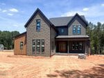 A modern two-story house at 150 Cherry Bark Drive, Youngsville, features dark brick and siding, large windows, and a covered front porch. It's set on a bare dirt lot with trees in the background under a blue sky.