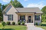 Nestled at 304 E Mason Street in Franklinton, this small beige house features dark shutters, a welcoming front porch with two chairs and potted plants, tidy shrubs, a gravel path to the black door, all surrounded by lush grass and trees.