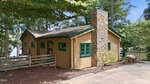 A small wooden cabin at 407 Sagamore Drive in Louisburg features tan siding, green-trimmed windows, and a stone chimney. Nestled among trees near water, it boasts a gravel driveway and wooden fence out front.