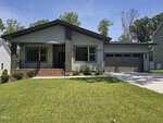 Single-story modern house at 113 Ottawa Drive, Louisburg, with gray siding, black trim, a covered front porch with railing, brick steps, large windows, a double garage, and a well-maintained green lawn under a blue sky.
